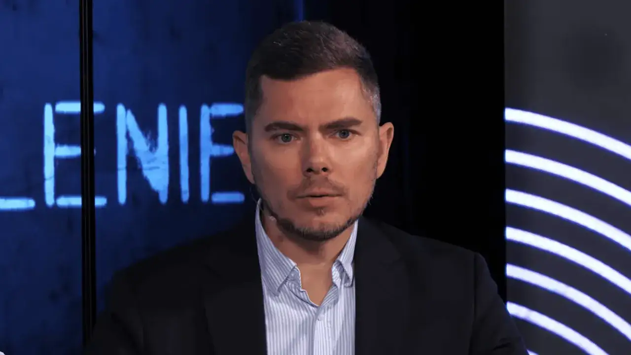 Portret Piotra Zychowicza w garniturze, na tle niebieskiego napisu.